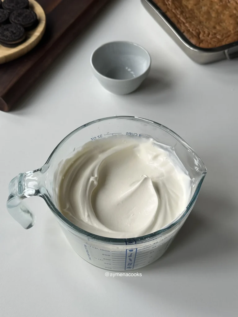 Thick, soft whipped cream in a glass measuring cup, ready for topping the cake.
