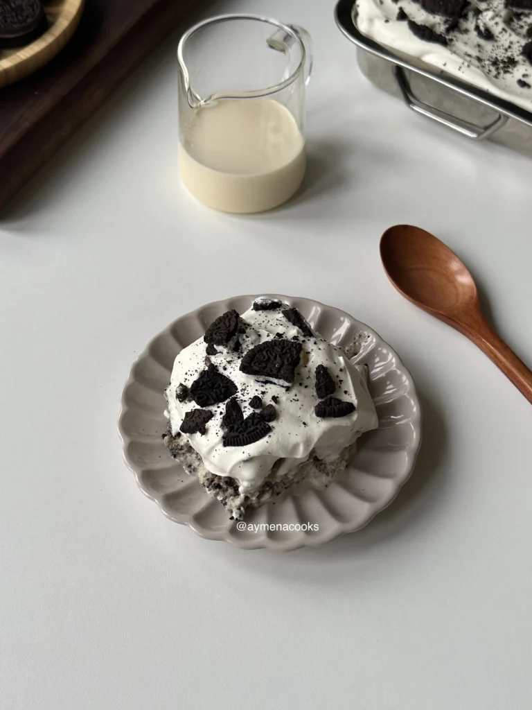 A slice of Oreo tres leches cake topped with whipped cream and crushed Oreos on a scalloped plate.