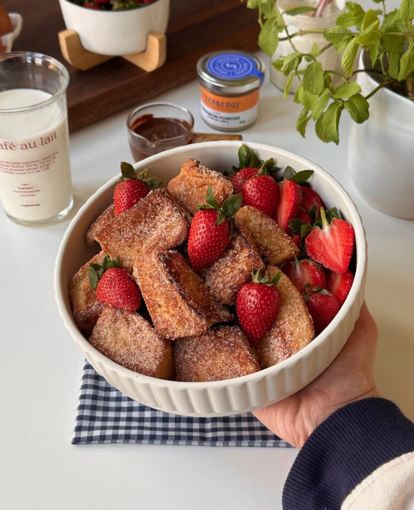 A hand holding a bowl of golden brioche French toast bites with fresh strawberries.