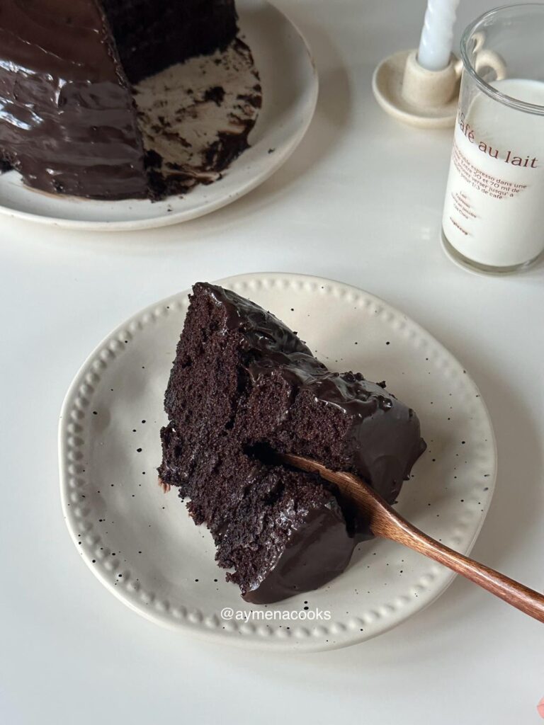 A fork going through a thick slice of three-layer Matilda chocolate cake covered in glossy fudge frosting.