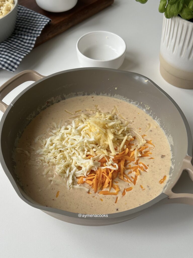 Three grated cheeses being added to stovetop mac and cheese sauce.