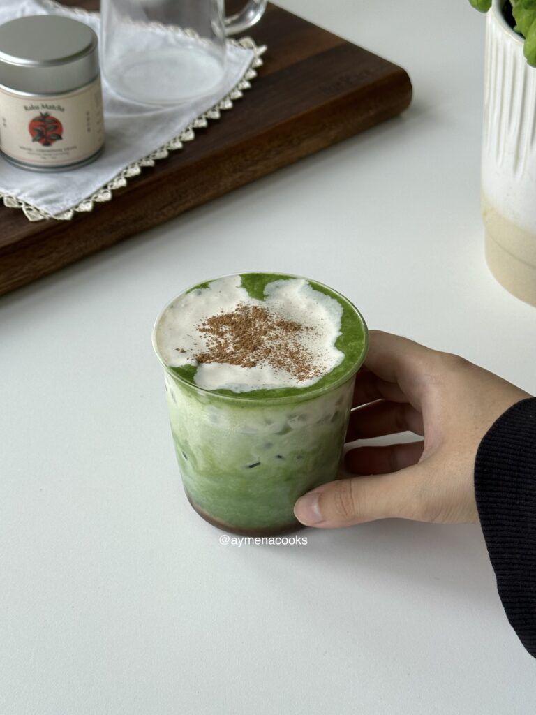 A hand holding a finished iced banana matcha with banana cold foam and cinnamon.