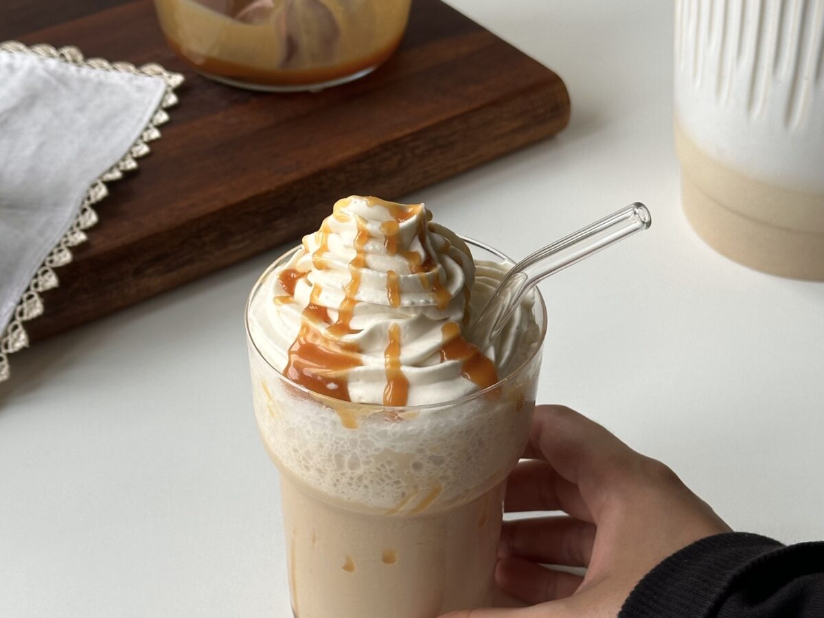 A hand holding a homemade caramel frappe topped with piped vanilla whipped cream and caramel sauce.