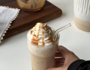 A hand holding a homemade caramel frappe topped with piped vanilla whipped cream and caramel sauce.