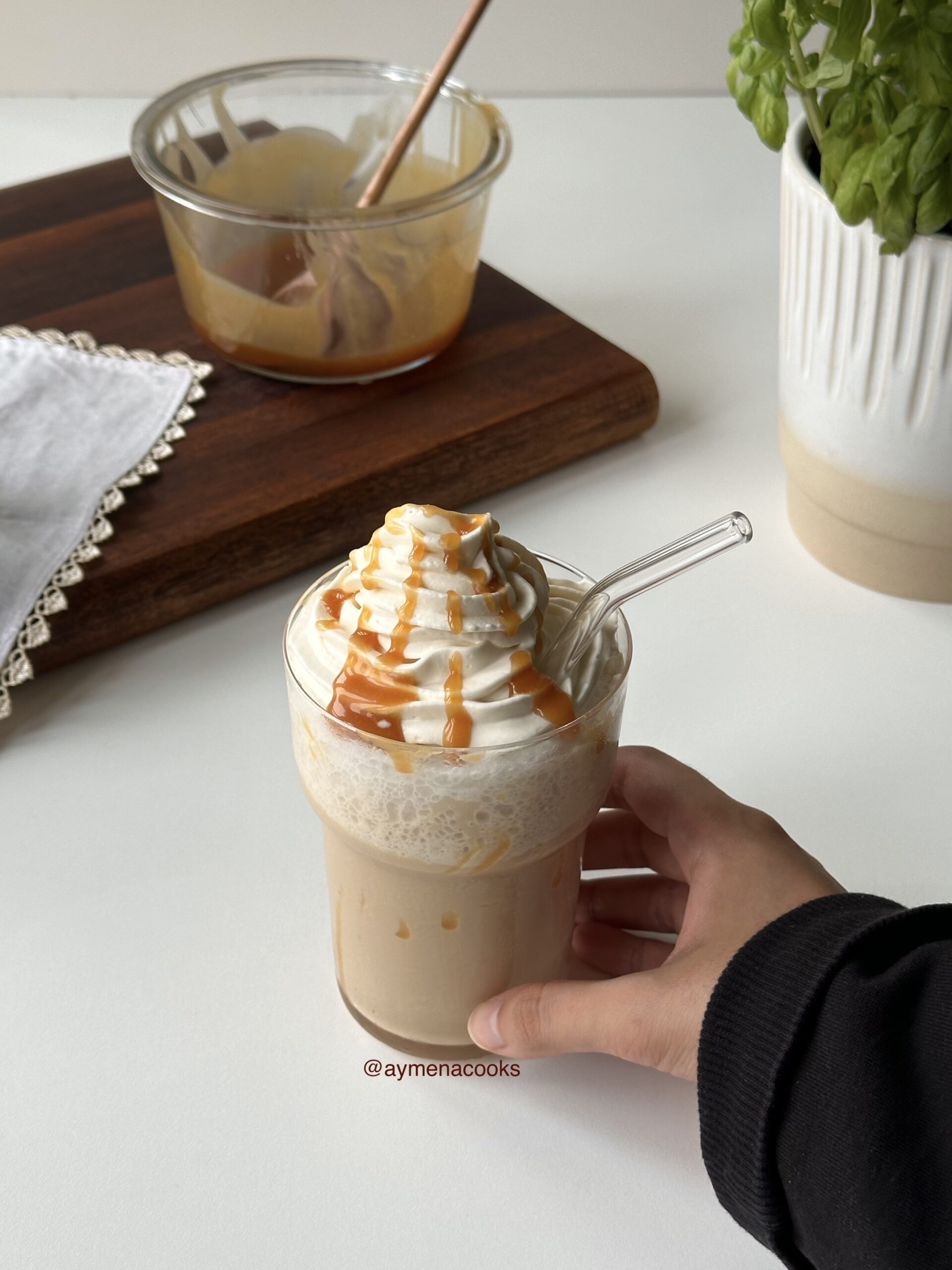 A hand holding a homemade caramel frappe topped with piped vanilla whipped cream and caramel sauce.