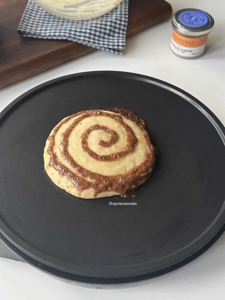 Cinnamon roll pancake cooking in the pan with bubbles on the surface and a melting brown sugar cinnamon swirl.
