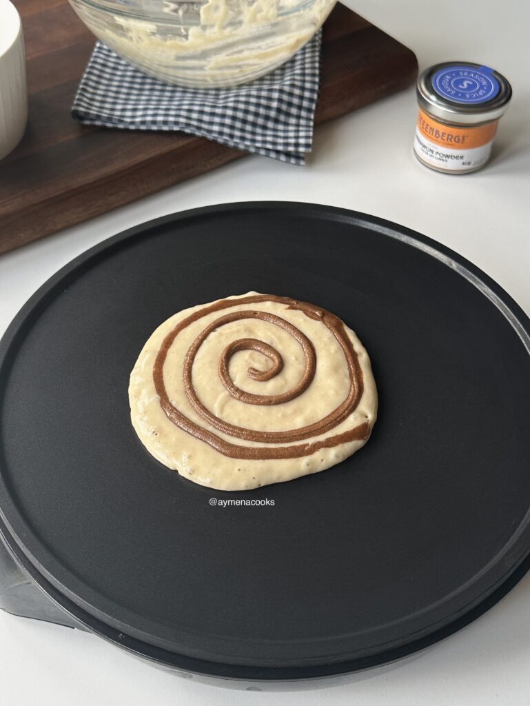 Cinnamon swirl piped over thick pancake batter being cooked in a nonstick pan.