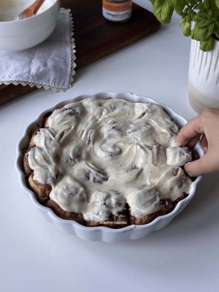 Brown butter cream cheese frosting spread over warm cinnamon roll bites and a hand pulling one of the bites out of the dish.