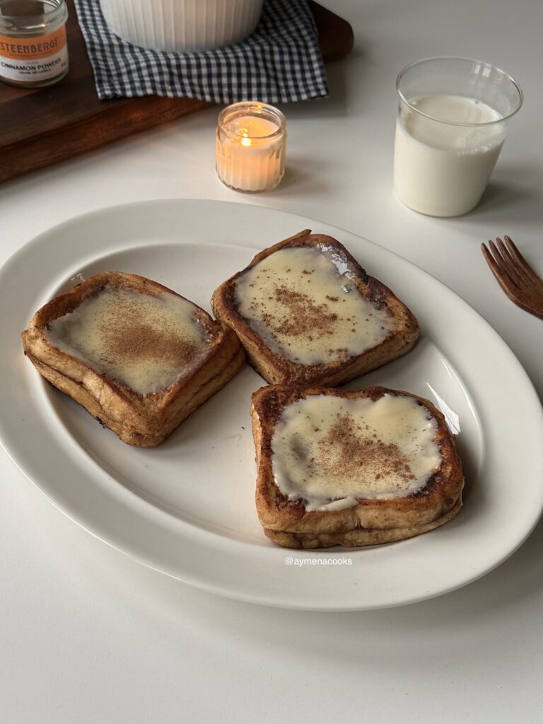 Three pieces of golden cinnamon roll French toast with cream cheese frosting on a serving platter.
